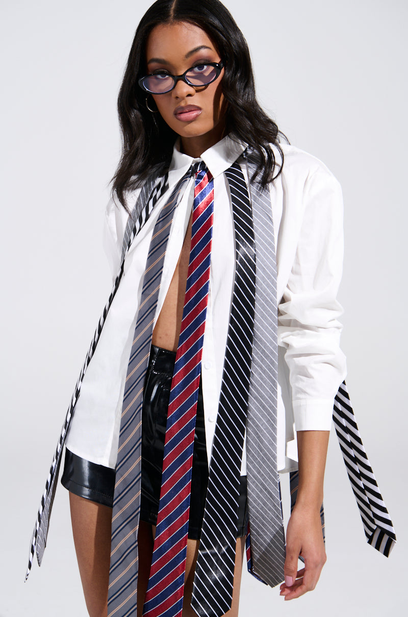 A woman with long dark hair and glasses wears the OH J SPECIAL TIE TOP, a white business-casual shirt adorned with cascading colorful ties. Paired with black shorts, her outfit stands out vividly against the minimalistic background.