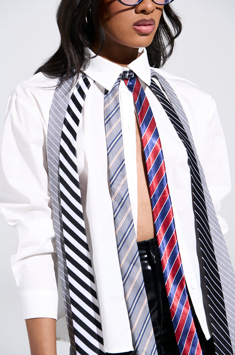 A person in a white button-up shirt highlights the OH J SPECIAL TIE TOP's longline silhouette with cascading ties in black, gray, blue, and red stripes. It complements their business-casual style while they peer through glasses, partially obscuring their face.