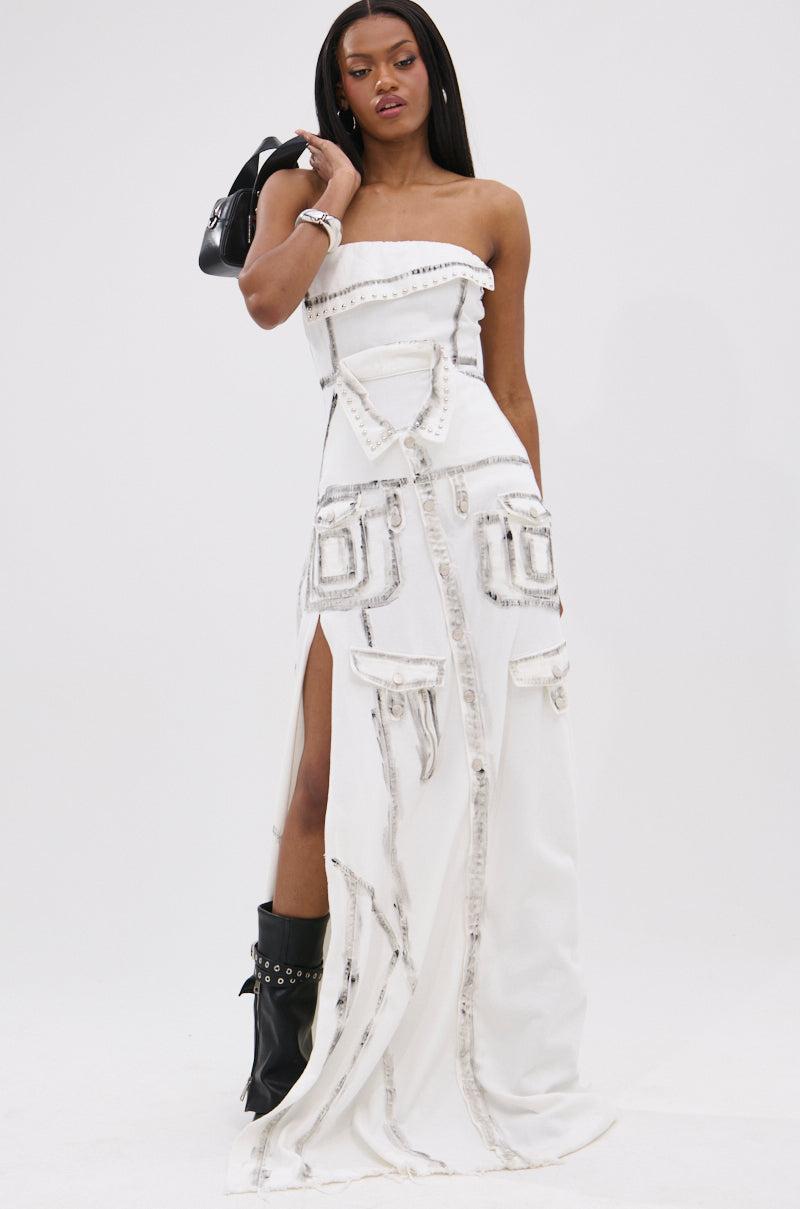 A woman stands confidently in the COVER STAR DENIM MAXI DRESS, a strapless white gown with textured rectangular details and front slits. She pairs it with a black handbag, black ankle boots, and a bracelet for an avant-garde look.