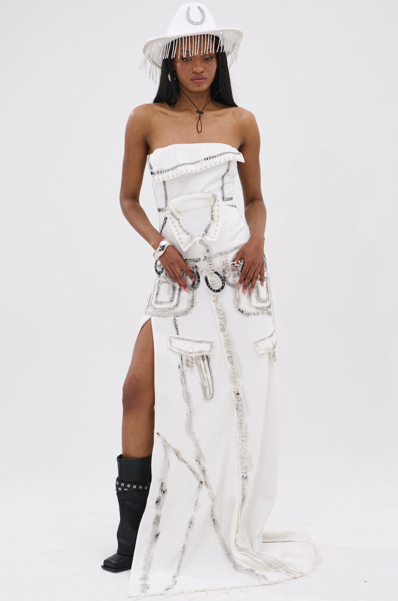 A woman wears the COVER STAR DENIM MAXI DRESS—an avant-garde, strapless white gown with rough-edged seams and a high side slit—styled with a white fringe cowboy hat, horseshoe emblem, black boots, and jewelry against a plain background.