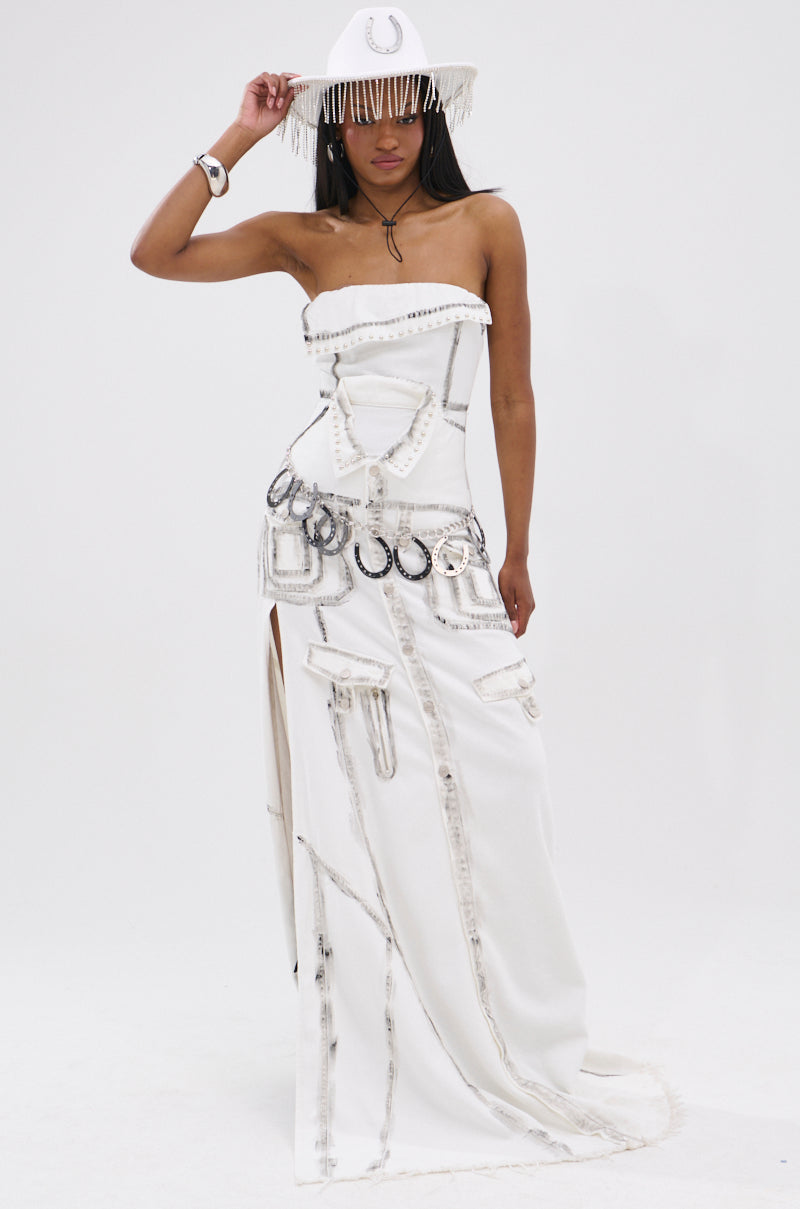 A woman wears the COVER STAR DENIM MAXI DRESS, a strapless white gown with silver accents and horseshoe shapes. She pairs it with a fringed white hat featuring a silver horseshoe emblem, posing against a plain white background.