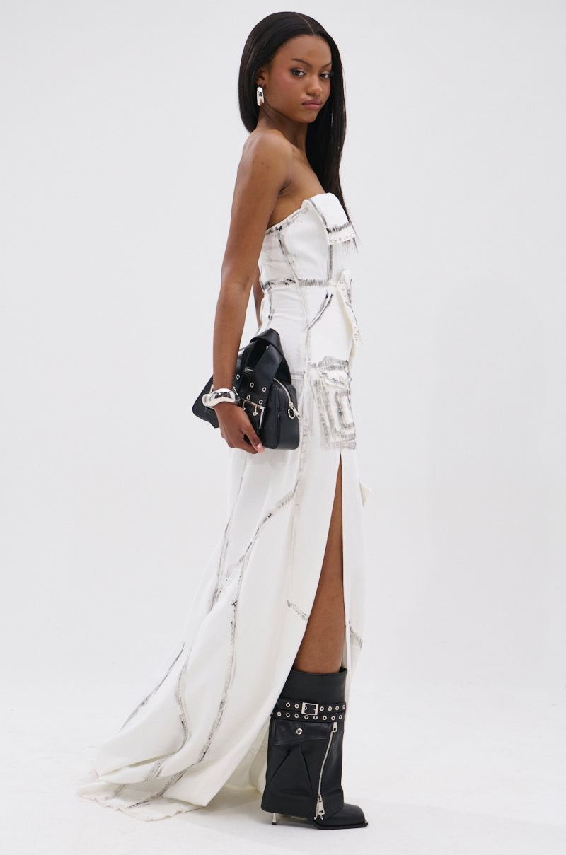 A woman wears the COVER STAR DENIM MAXI DRESS, a white strapless piece with silver accents and a thigh-high slit. She pairs it with a black handbag and black knee-high boots, her long straight hair set against a plain white background.