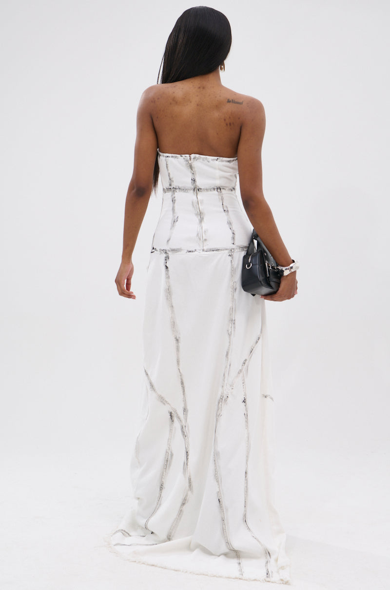 A woman with long dark hair stands facing away in the COVER STAR DENIM MAXI DRESS, a strapless white dress with subtle dark seams and an avant-garde silhouette. She holds a black handbag and wears bracelets, set against a plain white background.