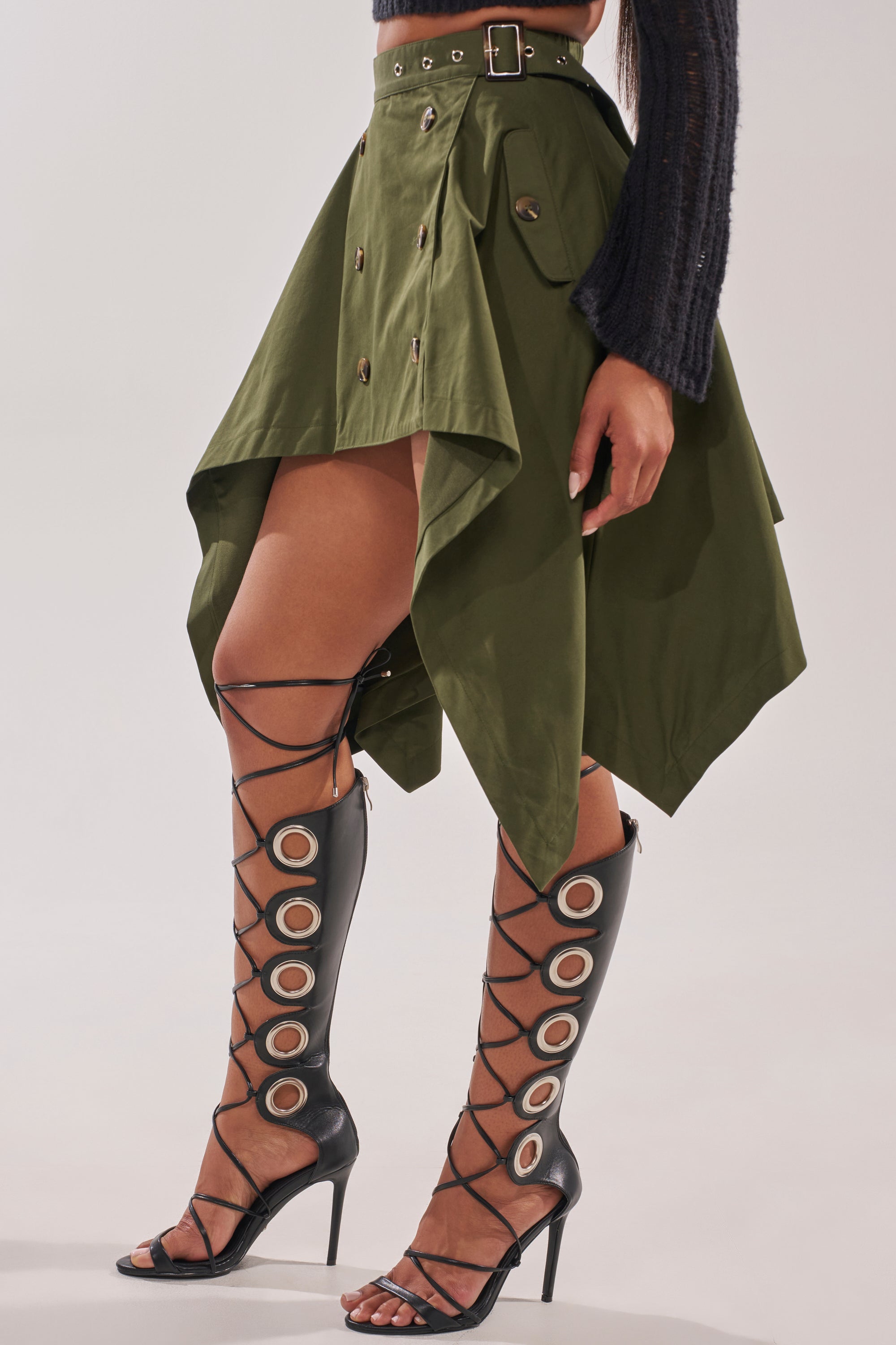 A woman wears the MAEVE MINI SKIRT IN OLIVE with buttons and a belt, styled with black, knee-high lace-up stiletto heels featuring large circular cutouts for a bold, streetwear-inspired look. 2