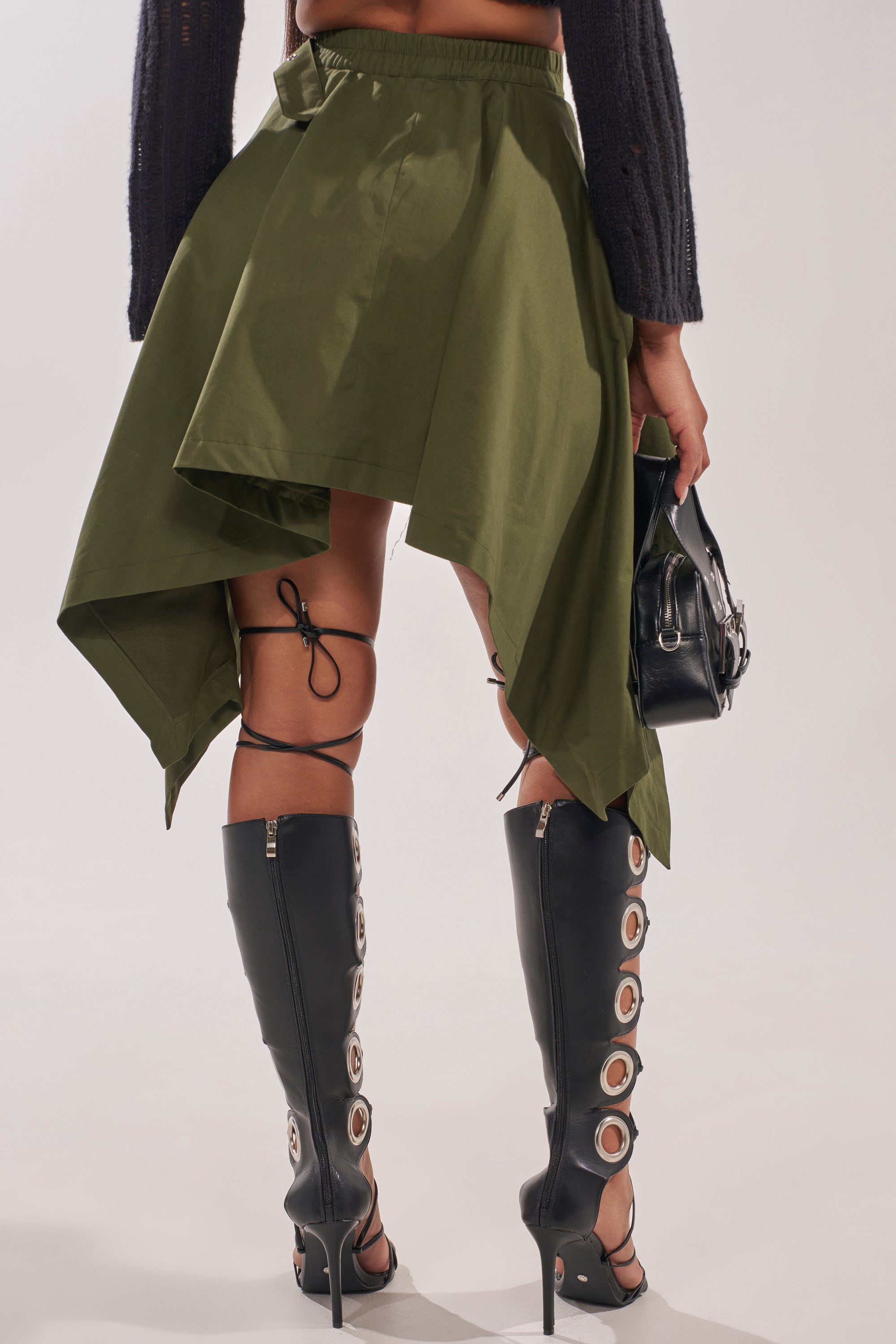 A person wearing the MAEVE MINI SKIRT IN OLIVE, black knee-high boots with circular cutouts and laces, and carrying a black handbag. The photo is taken from behind, showing only the lower half of their body. 3