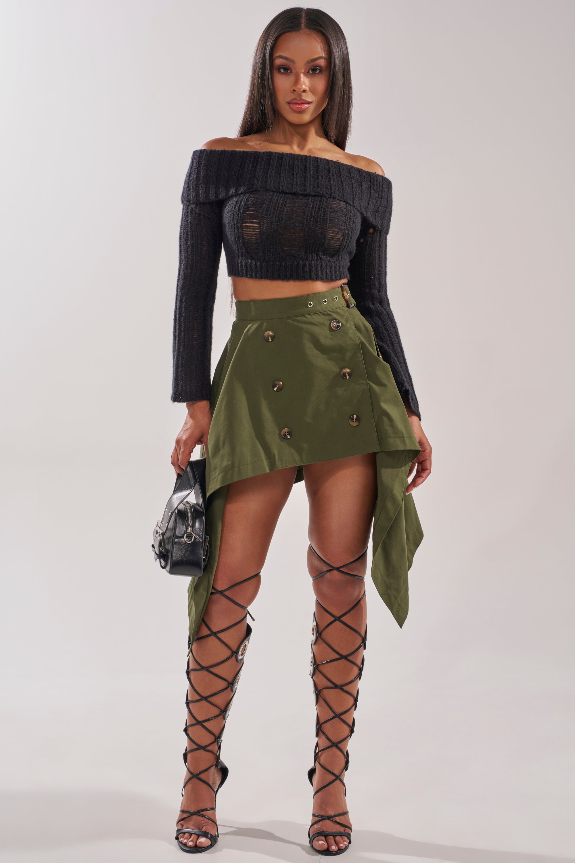 A woman stands facing forward, wearing a black off-shoulder cropped sweater, the MAEVE MINI SKIRT IN OLIVE with a handkerchief hem and buttons, tall black lace-up sandals, and holding a small black handbag by her side. 4