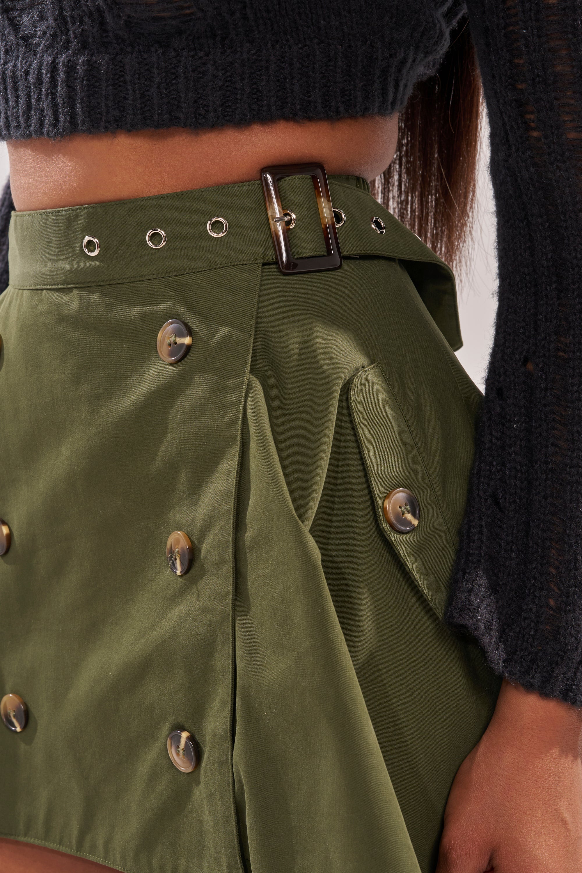 A close-up of a person wearing the MAEVE MINI SKIRT IN OLIVE, featuring large buttons and a square-buckle belt, styled with a black knit long-sleeve top for a chic, modern streetwear look. 5