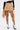 A person wearing the MAEVE MINI SKIRT in tan with gold buttons, a sheer black long-sleeve top, white socks with navy stripes, and black pointed heels with gold embellishments, shown from the waist down.
