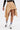 A person wears the MAEVE MINI SKIRT in beige with gold buttons, a black long-sleeve top, white socks with blue stripes, and black pointed heels with gold accents, standing against a plain white background.