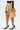 A person wearing the MAEVE MINI SKIRT in tan with button details, a black sheer long-sleeve top, white socks with blue stripes, and black pointed high heels with gold accents, standing against a plain white background.