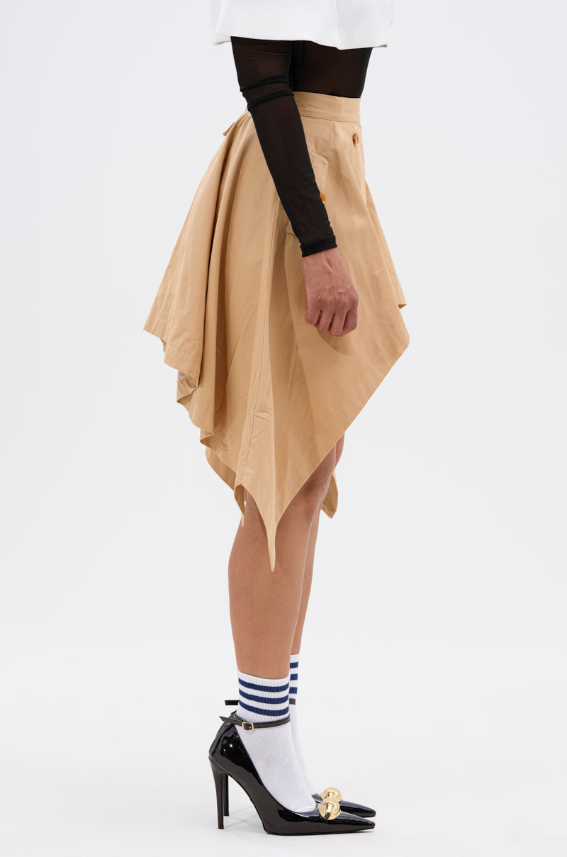 A person in profile wears the MAEVE MINI SKIRT in tan, styled with black mesh long sleeves, white socks featuring blue stripes, and black high-heeled shoes with gold accents against a plain white background.