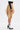 A person in profile wears the MAEVE MINI SKIRT in tan, styled with black mesh long sleeves, white socks featuring blue stripes, and black high-heeled shoes with gold accents against a plain white background.