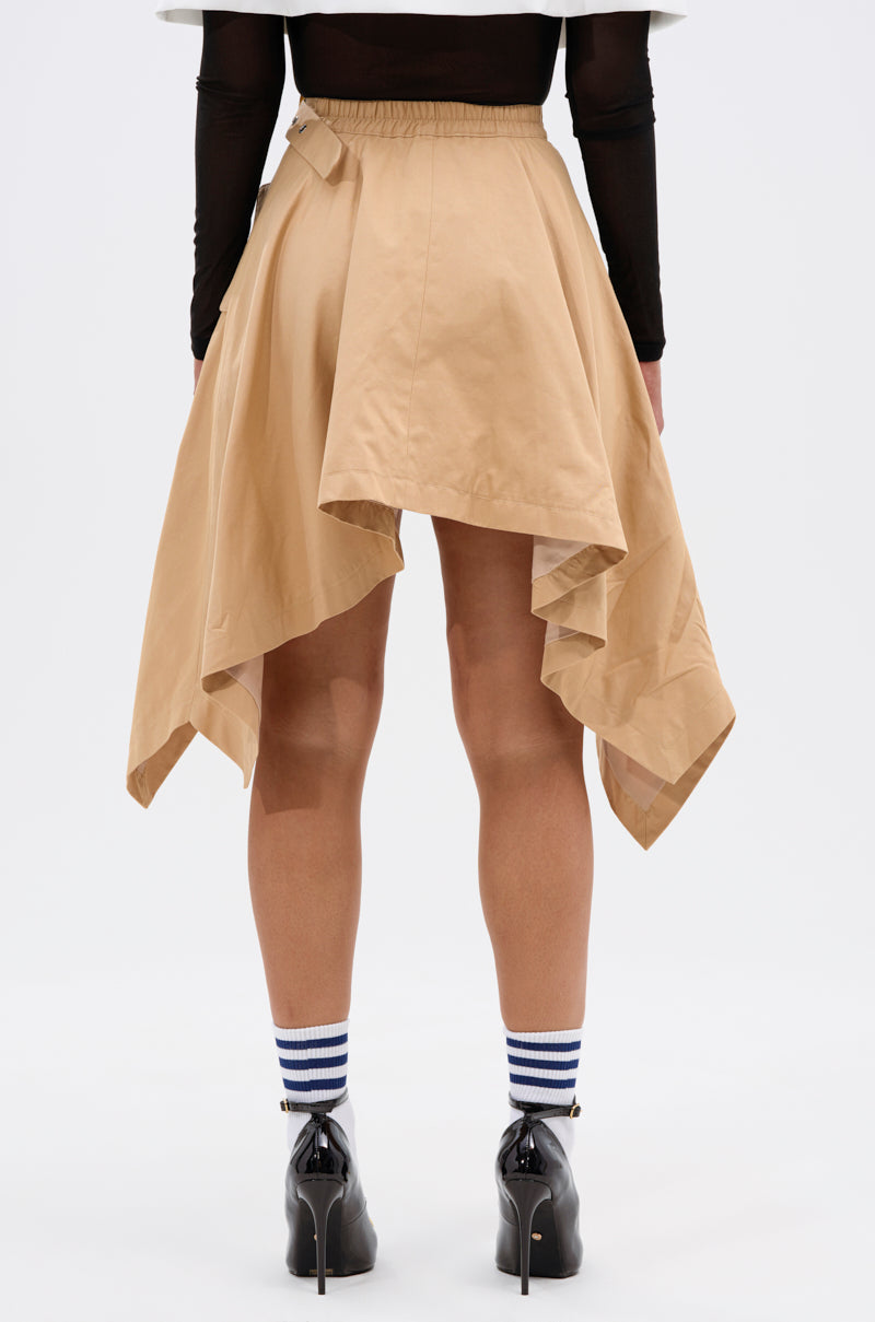A person stands facing away, wearing the MAEVE MINI SKIRT—a tan asymmetric skirt with layered, uneven hems—paired with a black long-sleeve top, black heeled shoes, and white crew socks with blue stripes. The background is plain white.