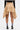 A person stands facing away, wearing the MAEVE MINI SKIRT—a tan asymmetric skirt with layered, uneven hems—paired with a black long-sleeve top, black heeled shoes, and white crew socks with blue stripes. The background is plain white.