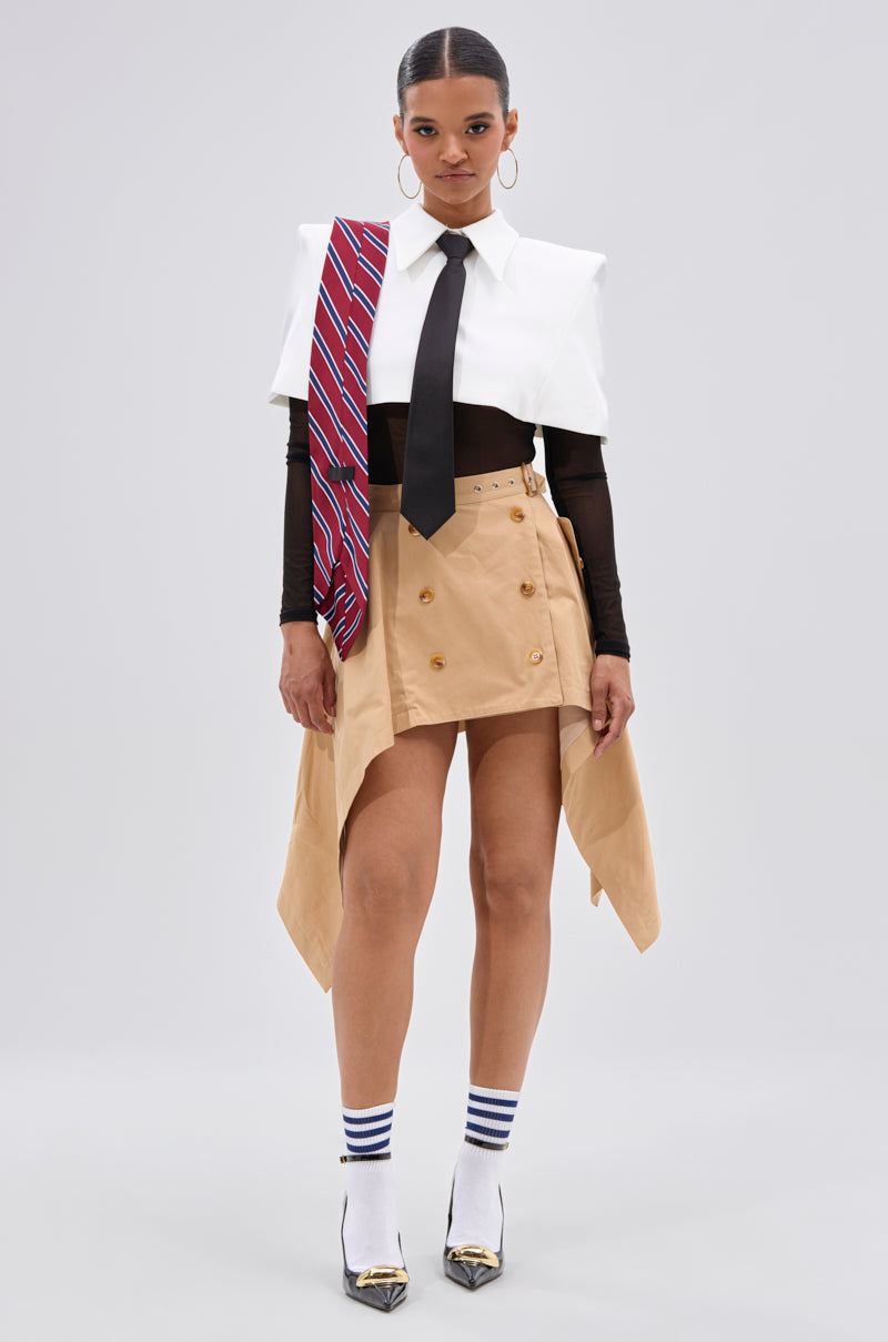 A woman faces forward wearing a white cropped shirt with padded shoulders, a black tie, a red striped tie over her shoulder, the MAEVE MINI SKIRT, white striped socks, and pointed heels.