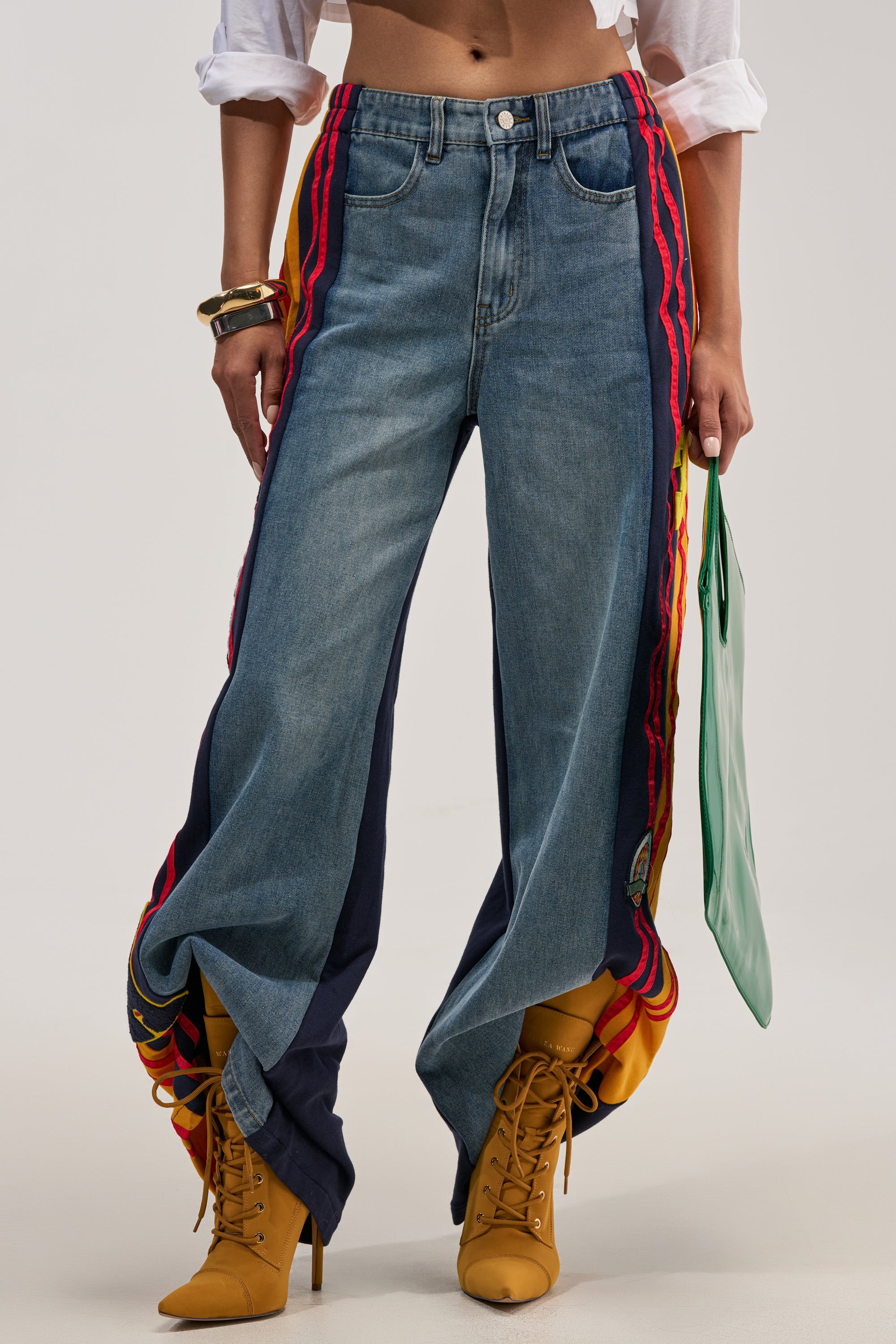 Wearing the LIGHTS OUT TWO-IN-ONE JEANS—sporty wide-leg blue denim featuring colorful side panels—paired with a white shirt, yellow lace-up boots, gold bangles, and a mint green clutch. The long track pants bunch at the ankles, bringing a playful vibe to this varsity-inspired streetwear ensemble.