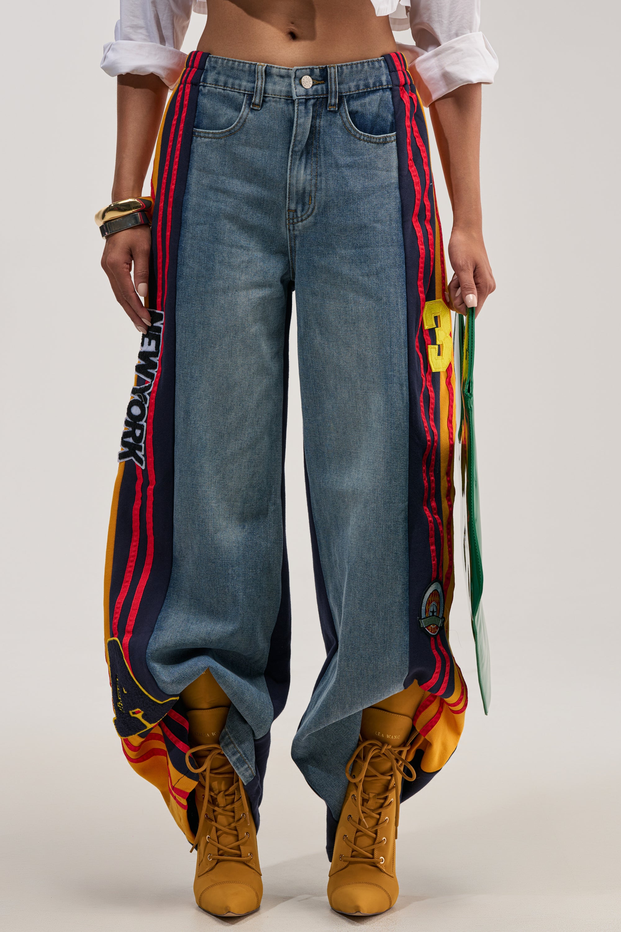 A person wearing the LIGHTS OUT TWO-IN-ONE JEANS, styled with colorful side panels and bold "NEW YORK" varsity-inspired graphics, paired with yellow boots, rolled-up sleeves, stacked bracelets, and carrying a green and white bag for a standout streetwear look.
