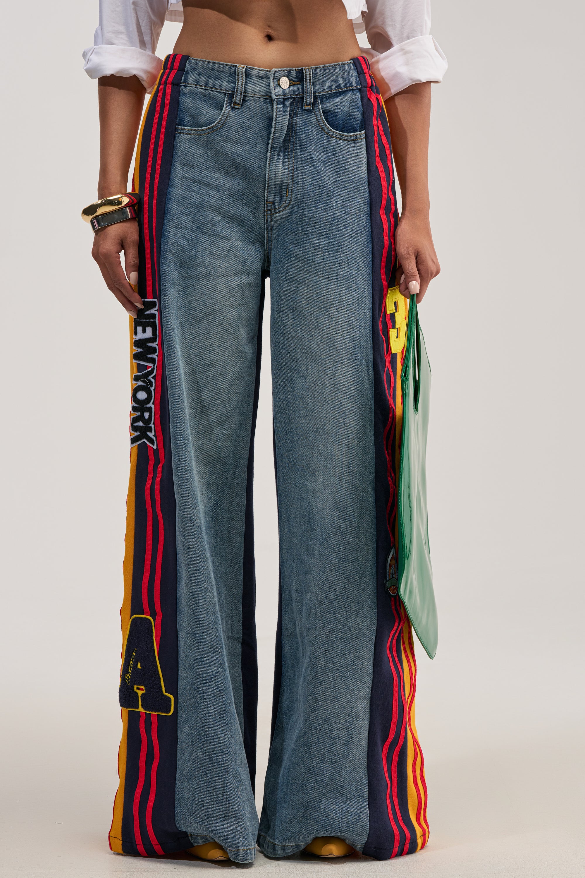 Person wearing the LIGHTS OUT TWO-IN-ONE JEANS—varsity-inspired streetwear featuring sporty wide-leg denim track pants with bold red, yellow, and navy side stripes, graphic patches including “NEW YORK” and an “A”—holding a green bag, with rolled-up white shirt sleeves visible.