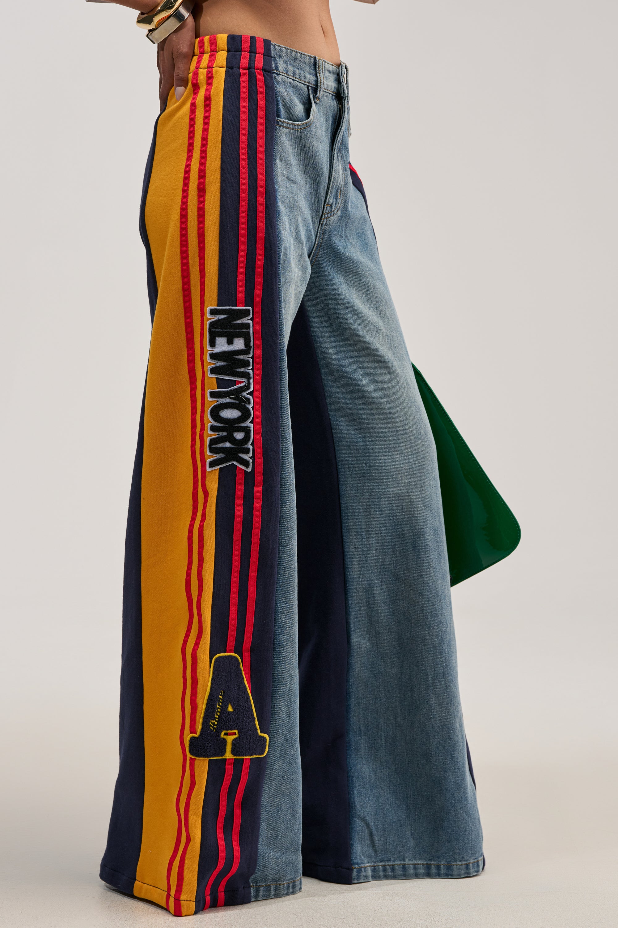 A person wearing the LIGHTS OUT TWO-IN-ONE JEANS—denim track pants featuring yellow and red striped side panels, "NETWORK" text detailing, and a bold varsity-style "A" patch—captures varsity-inspired streetwear flair. They are holding a green bag, with only their lower torso and legs visible.