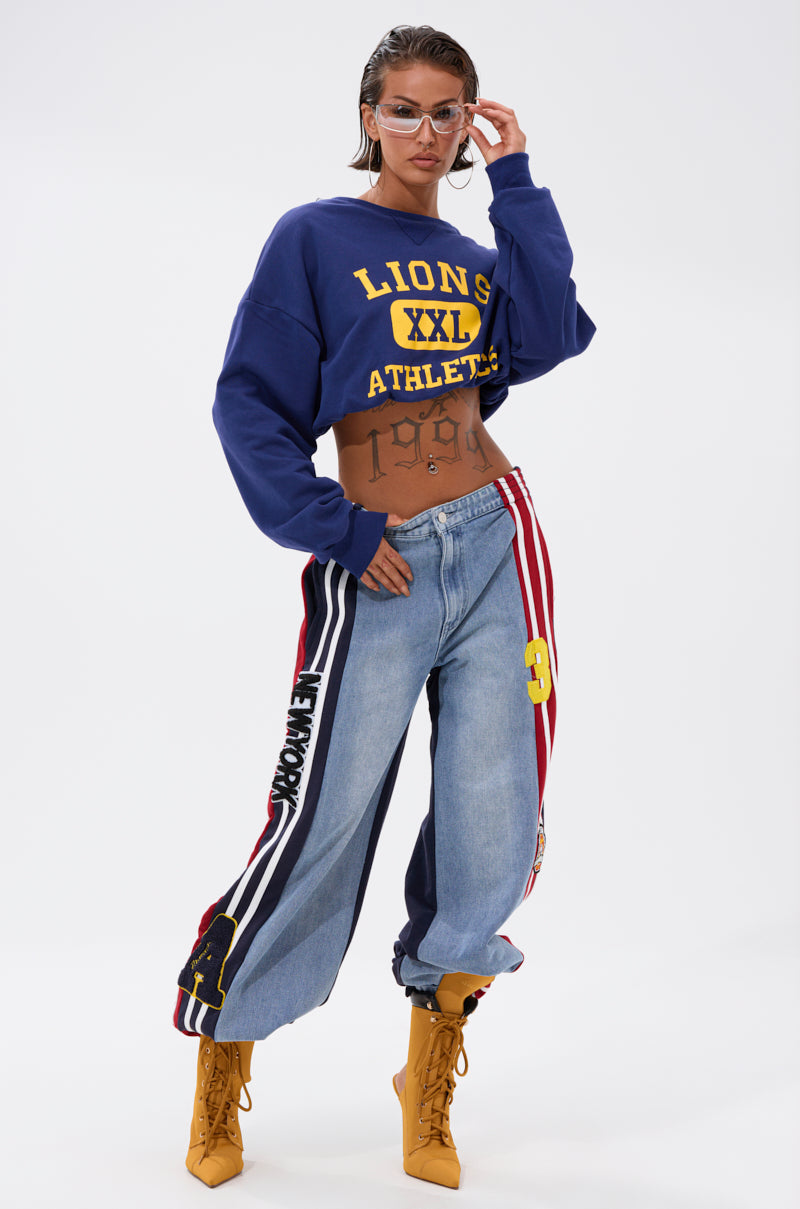 A woman poses confidently in a blue "LIONS XXL ATHLETICS" crop sweatshirt, LIGHTS OUT TWO-IN-ONE JEANS with wide-leg patches and stripes, yellow lace-up boots, clear-framed glasses, wet-look hair, and visible tattoos.