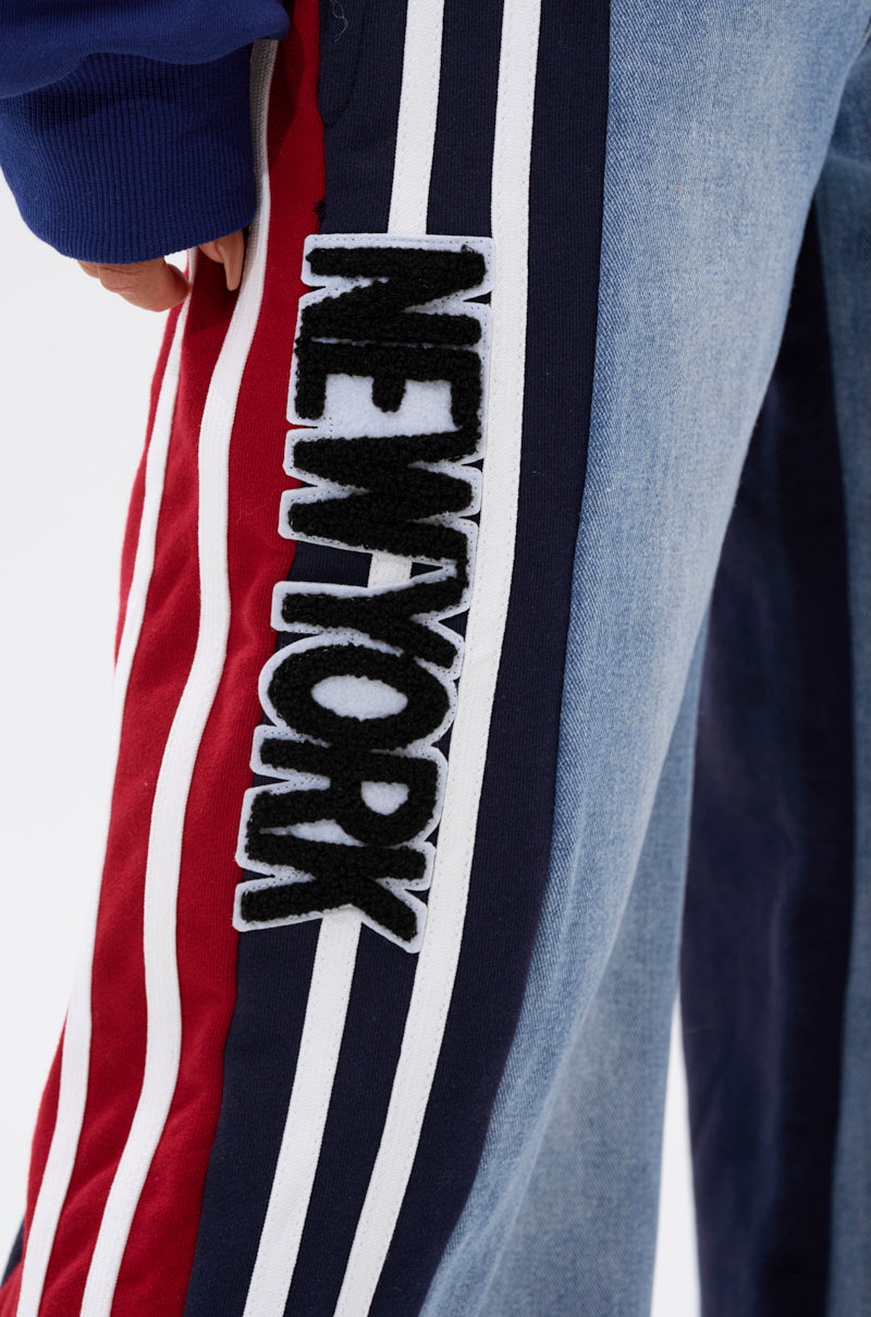 A close-up of a person wearing LIGHTS OUT TWO-IN-ONE JEANS, featuring a wide-leg silhouette, red, white, and navy side stripes, and "NEW YORK" in bold black-and-white letters sewn onto the side.