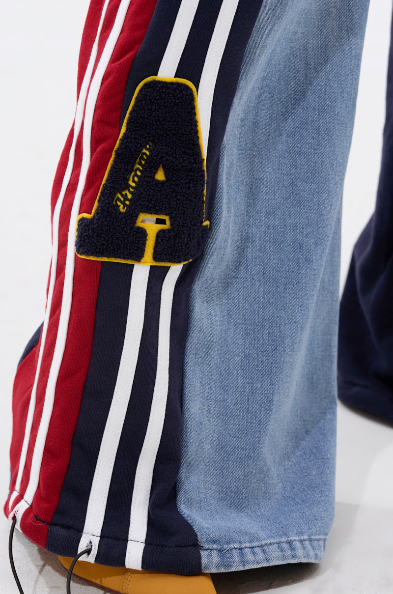 Close-up of the LIGHTS OUT TWO-IN-ONE JEANS, featuring hybrid denim with red, white, and blue stripes and light blue panels. A bold black "A" patch outlined in yellow accents the wide-leg silhouette for a sporty finish.