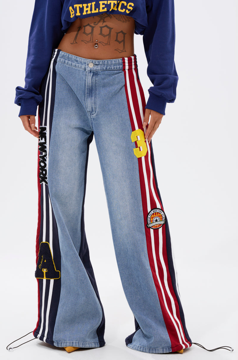 Wearing the LIGHTS OUT TWO-IN-ONE JEANS—hybrid patchwork jeans with red and white stripes, varsity patches, and "NEW YORK" text—paired with a cropped “ATHLETICS 1999” top that reveals a tattooed midriff.