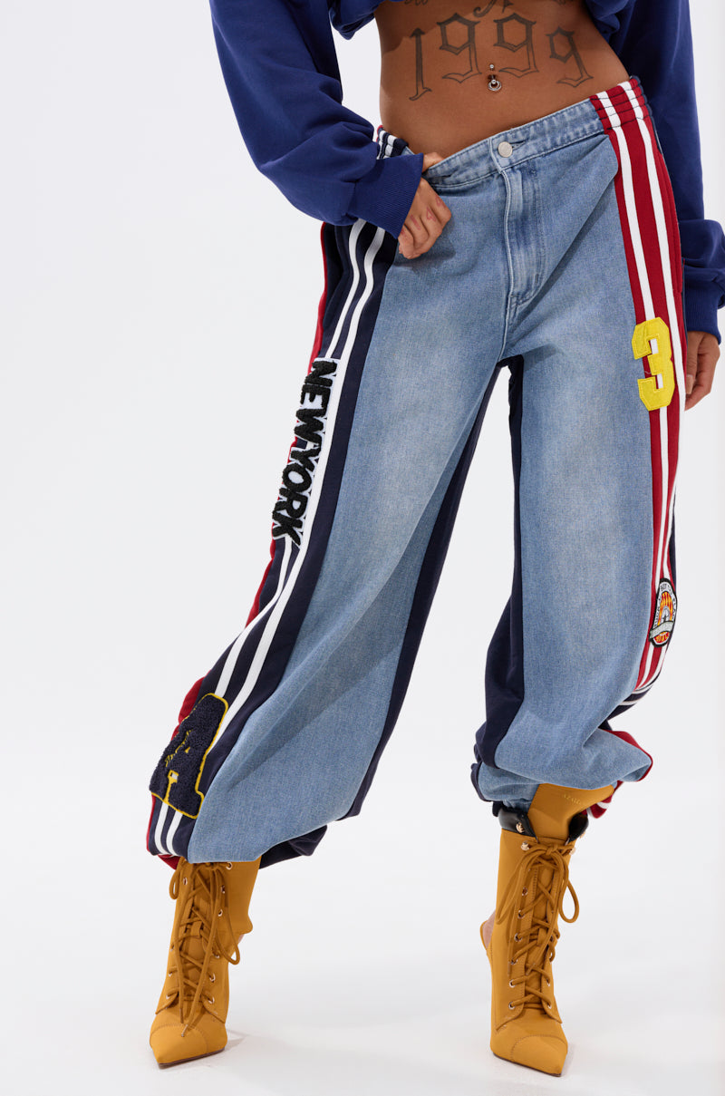 A person in LIGHTS OUT TWO-IN-ONE JEANS featuring a wide-leg patchwork design with "NEW YORK" text, stripes, and a yellow "3," paired with a cropped blue top showing a "1999" tattoo and yellow lace-up boots.