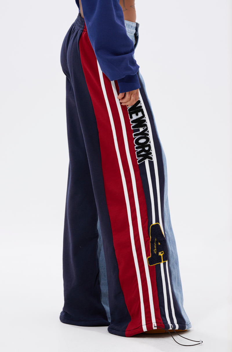 Side view of a person wearing the LIGHTS OUT TWO-IN-ONE JEANS featuring wide-leg hybrid design, red, navy, and white stripes, “NEW YORK” text, and a yellow patch. Styled with a blue sweatshirt for a standout sporty look.