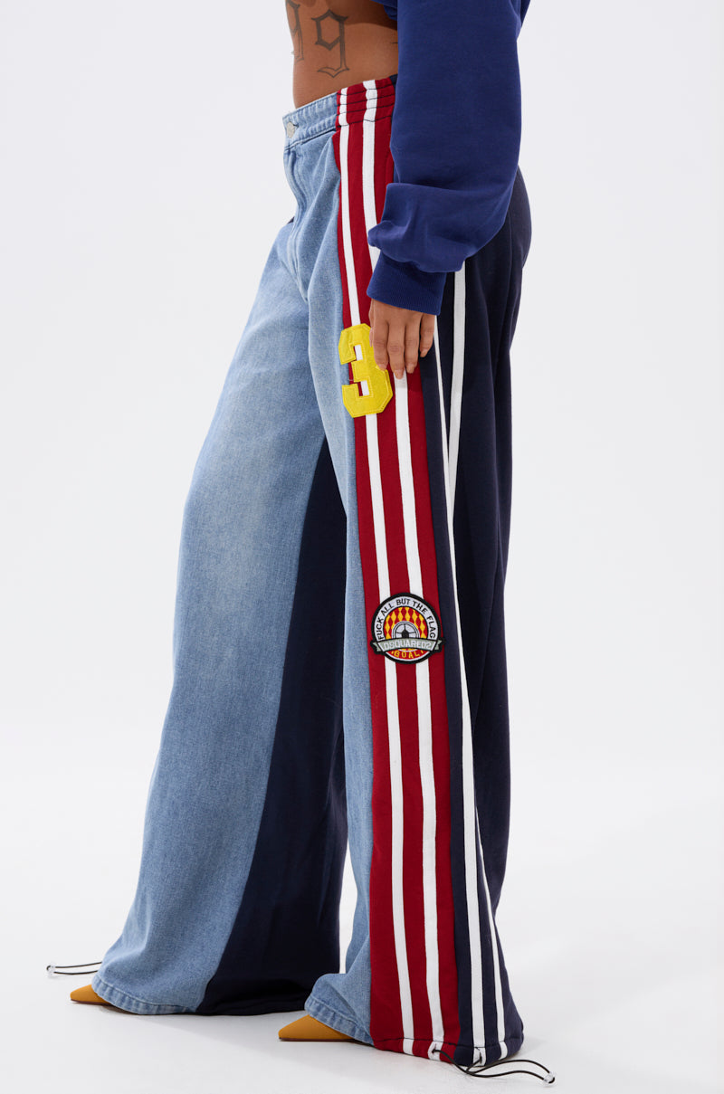 A person wears the LIGHTS OUT TWO-IN-ONE JEANS—hybrid wide-leg pants featuring light blue denim on one side and navy with red/white stripes, a yellow “3” patch, and round embroidery—styled with a blue sweatshirt and yellow pointed shoes.