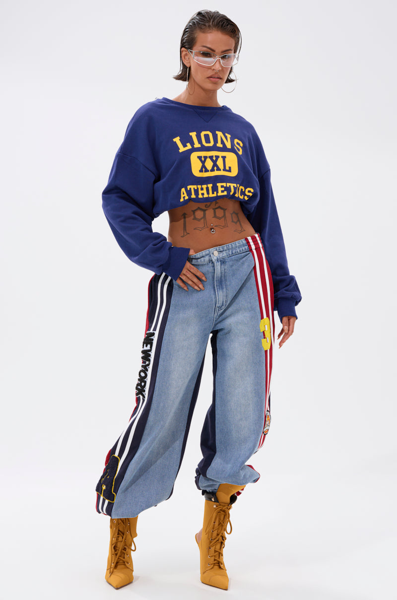 A woman poses confidently in a cropped blue "LIONS XXL ATHLETICS" sweatshirt, LIGHTS OUT TWO-IN-ONE JEANS with colorful patches, yellow lace-up boots, and clear-framed glasses. She sports wet-look hair and minimal jewelry.