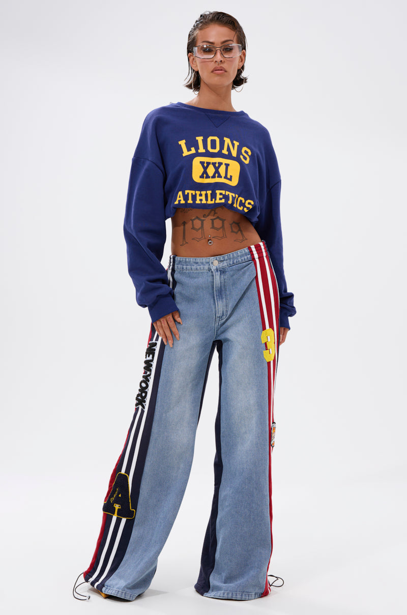 A person wears sunglasses, a blue cropped "Lions XXL Athletics" sweatshirt, and LIGHTS OUT TWO-IN-ONE JEANS with multicolored side stripes and patches, standing confidently with visible torso tattoos against a plain white background.