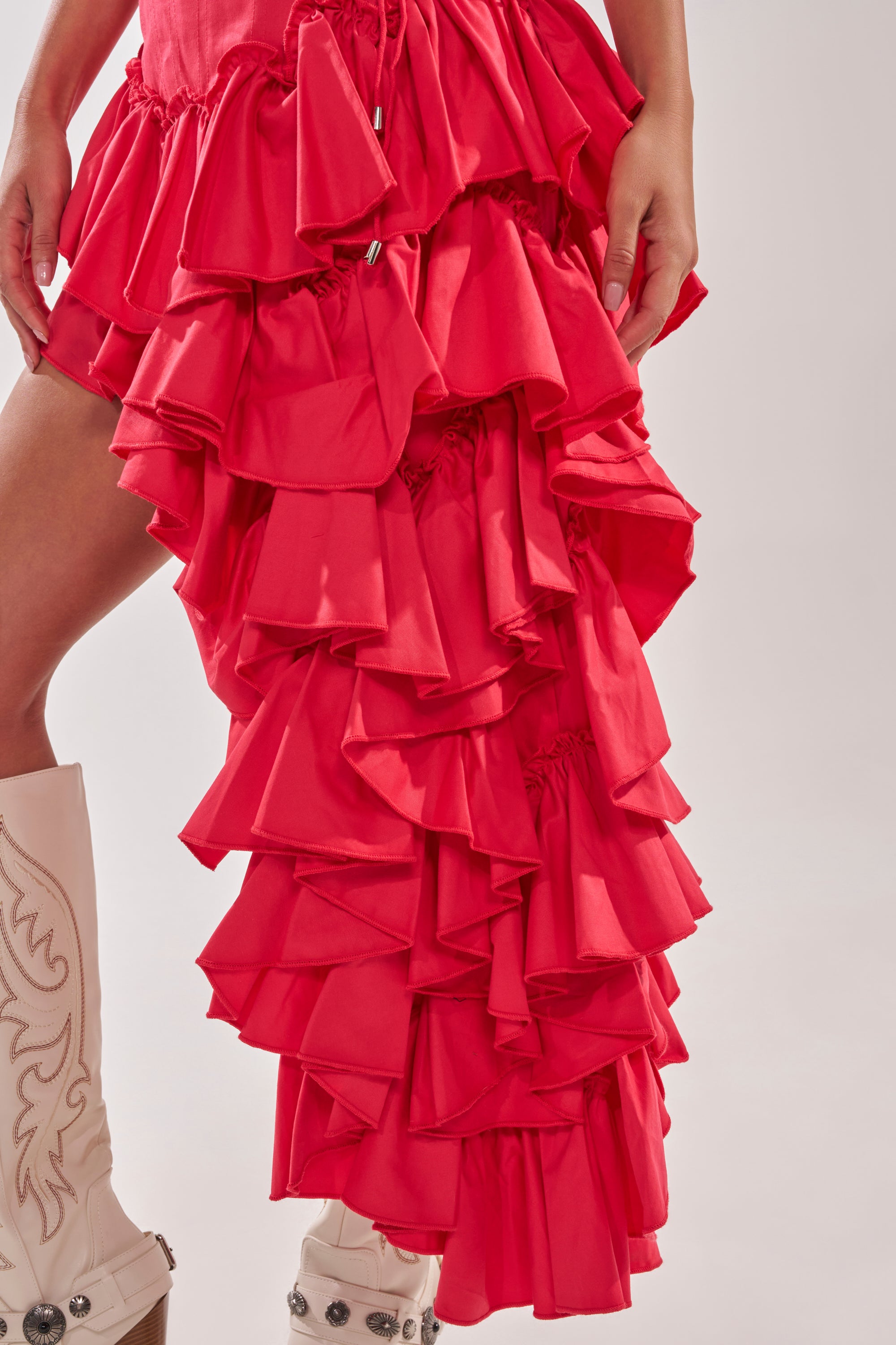 A person wears the CLAIRE HIGH LOW RUFFLE SKIRT IN RED and embroidered white cowboy boots, standing against a plain light background. 10