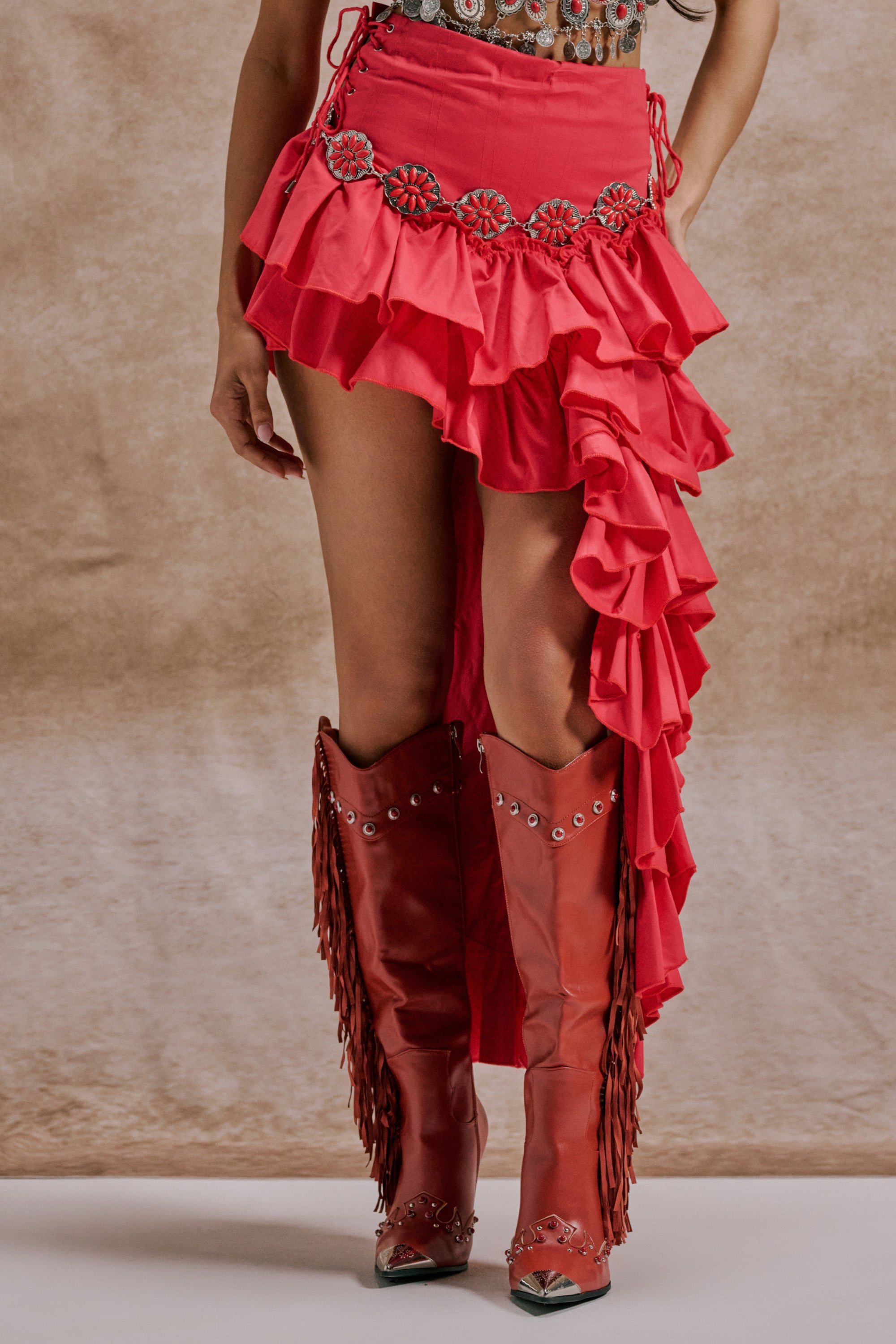 Wearing the CLAIRE HIGH LOW RUFFLE SKIRT IN RED and matching red fringe cowboy boots, a person stands against a beige background. 1