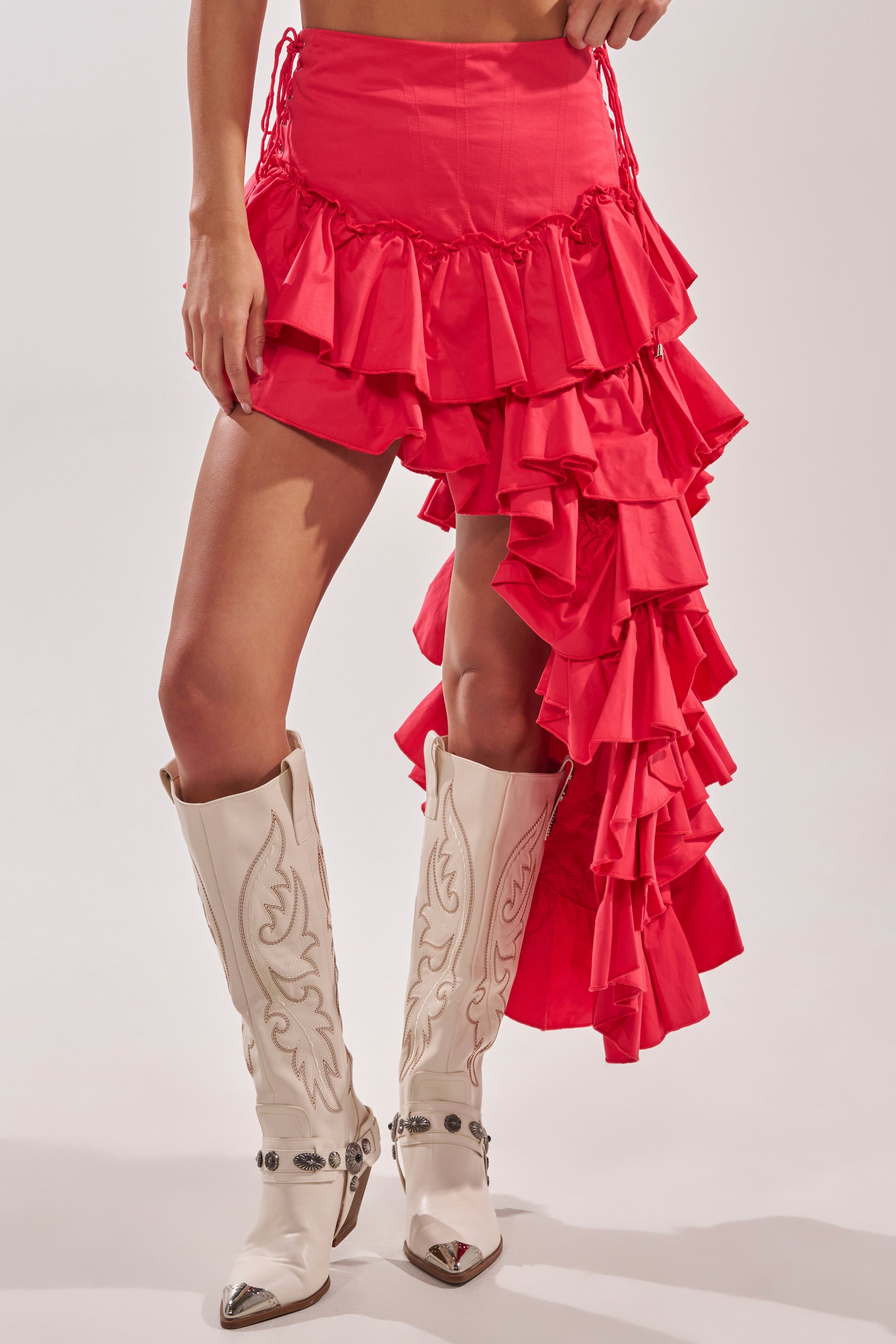 Wearing the CLAIRE HIGH LOW RUFFLE SKIRT IN RED featuring an asymmetrical hem and lace-up sides, paired with white embroidered cowboy boots. Only the lower half of the body is shown. 3
