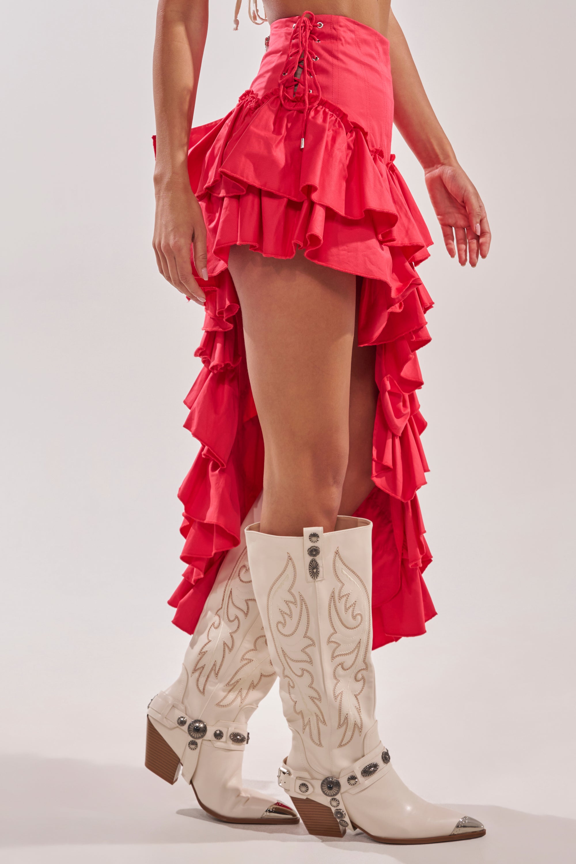 A person wears the CLAIRE HIGH LOW RUFFLE SKIRT IN RED with lace-up sides and white cowboy boots featuring decorative stitching and silver accents. Only the lower body is visible against a plain background. 4