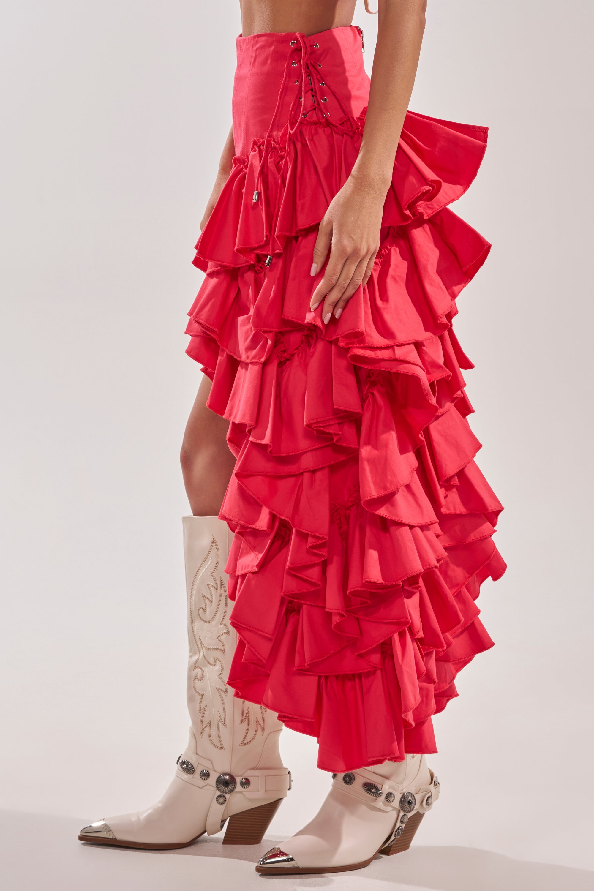 A person wears the CLAIRE HIGH LOW RUFFLE SKIRT IN RED, featuring a high waist and ruffle details, paired with white embroidered cowboy boots. Only the lower half of their body is visible. 5