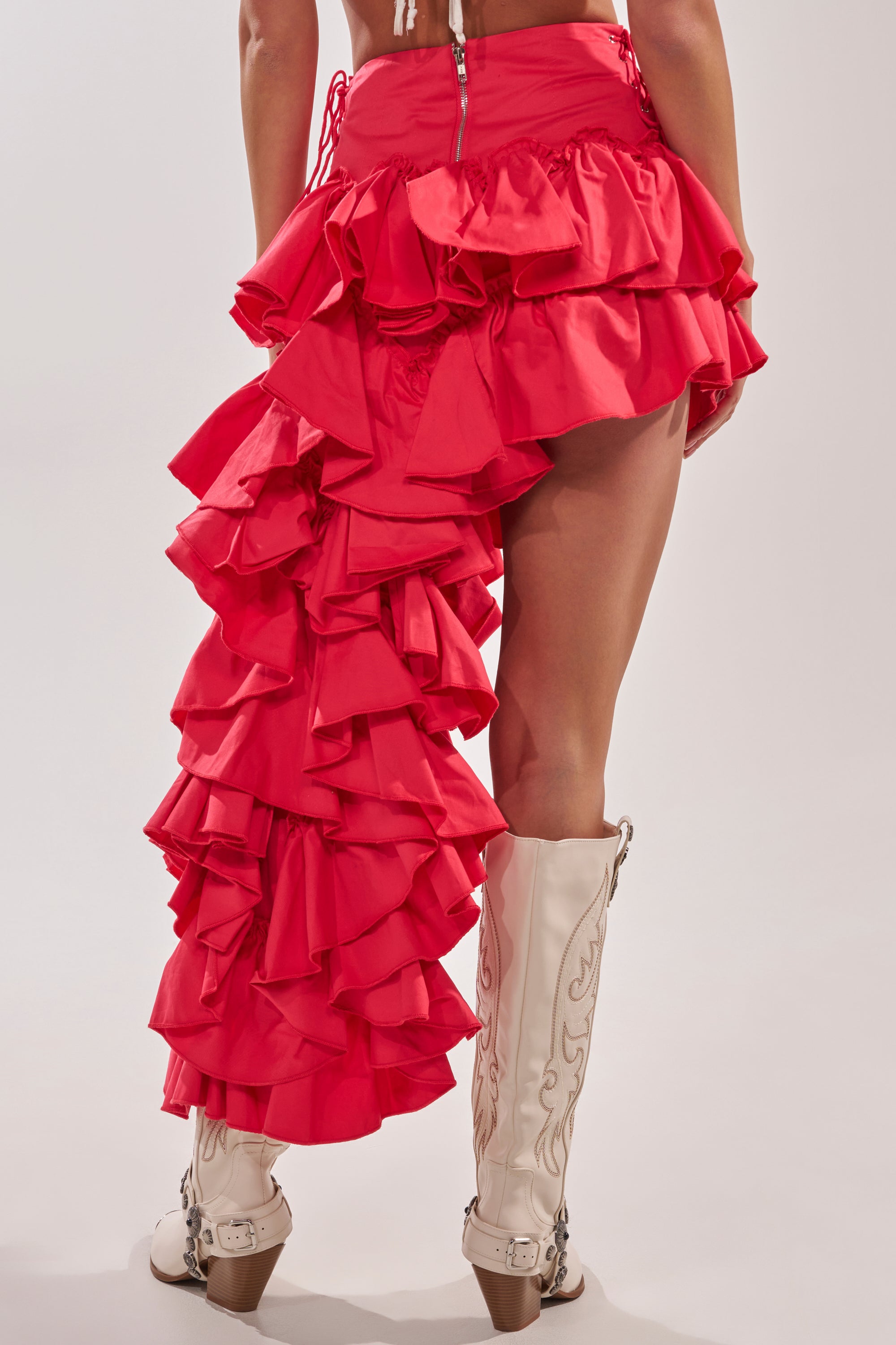 A person wears the CLAIRE HIGH LOW RUFFLE SKIRT IN RED, featuring an asymmetrical, layered ruffle design, paired with cream cowboy boots with decorative stitching. The background is plain and neutral. 6