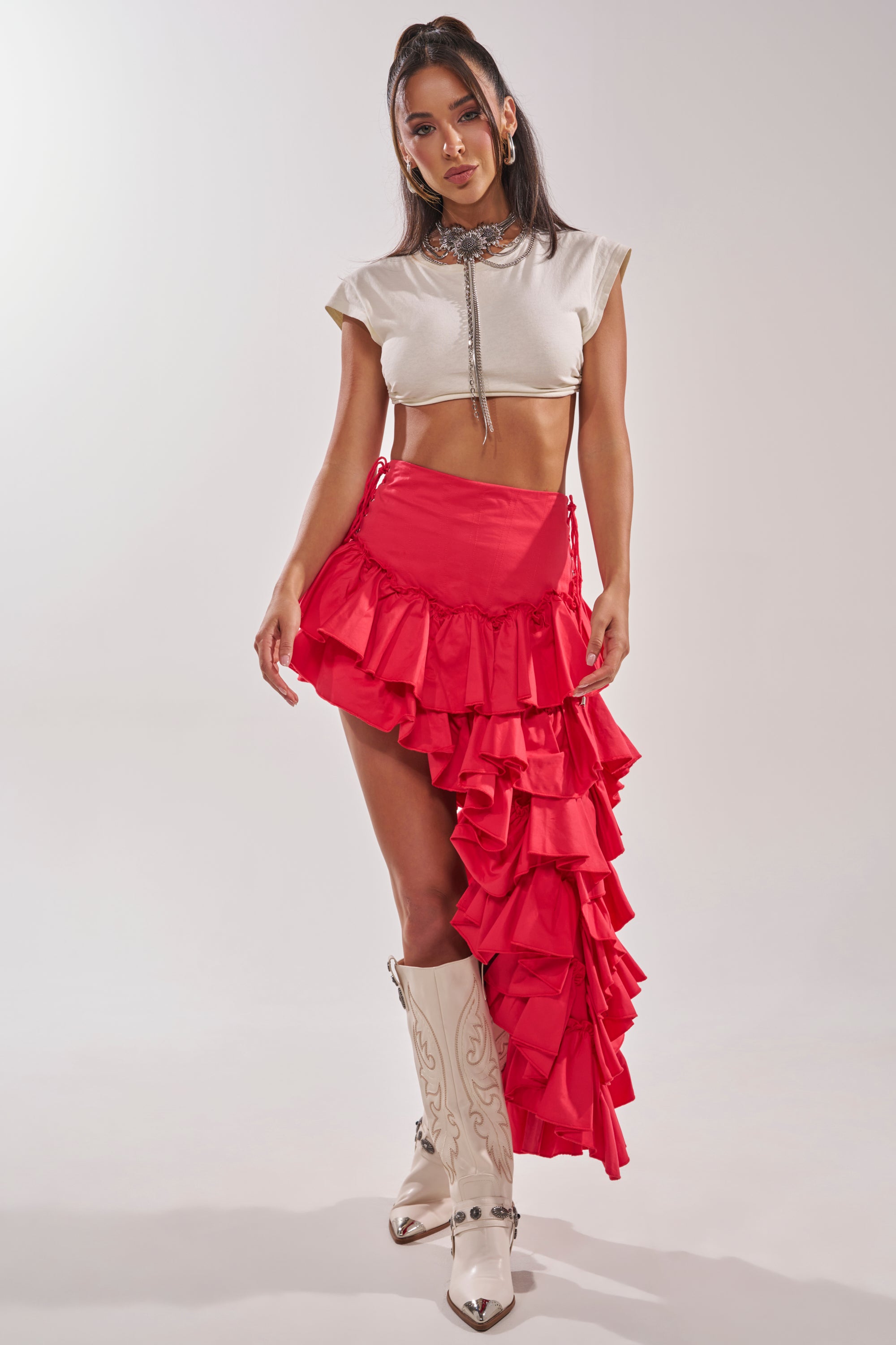 A woman faces forward against a plain background in a white crop top, the CLAIRE HIGH LOW RUFFLE SKIRT IN RED, and white cowboy boots, accessorized with earrings and a choker necklace. 8
