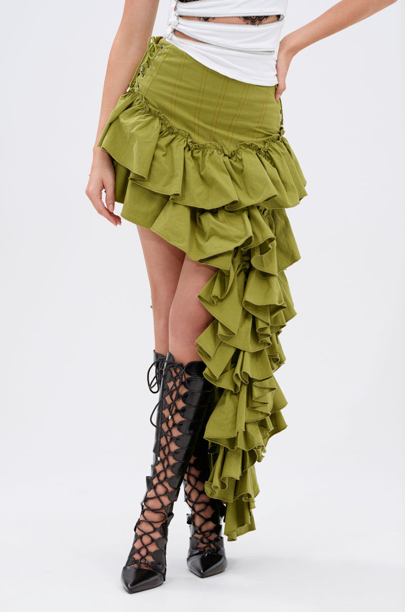 A person wears the CLAIRE HIGH LOW RUFFLE SKIRT in olive green with lace-up details, styled with a white top and black knee-high lace-up heeled boots. Only the lower body is shown against a plain background.