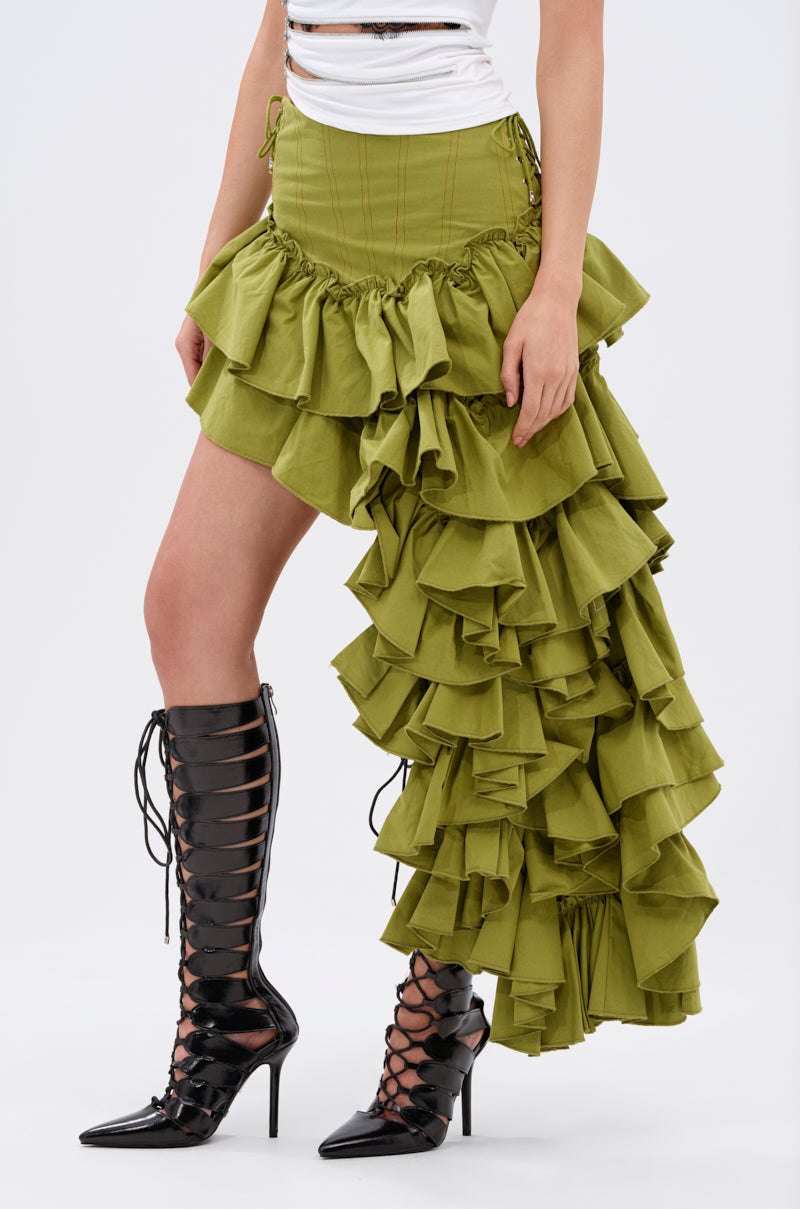 Wearing a white sleeveless top, the CLAIRE HIGH LOW RUFFLE SKIRT in olive green with long layered sides, and knee-high black lace-up heeled boots, a person stands against a plain background.