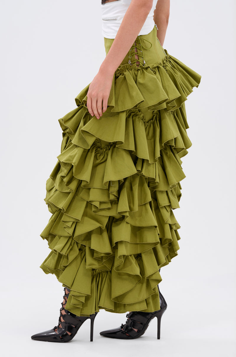 A person models the CLAIRE HIGH LOW RUFFLE SKIRT in olive green, styled with a white top and black pointed-toe heels, photographed from the side against a plain light background.