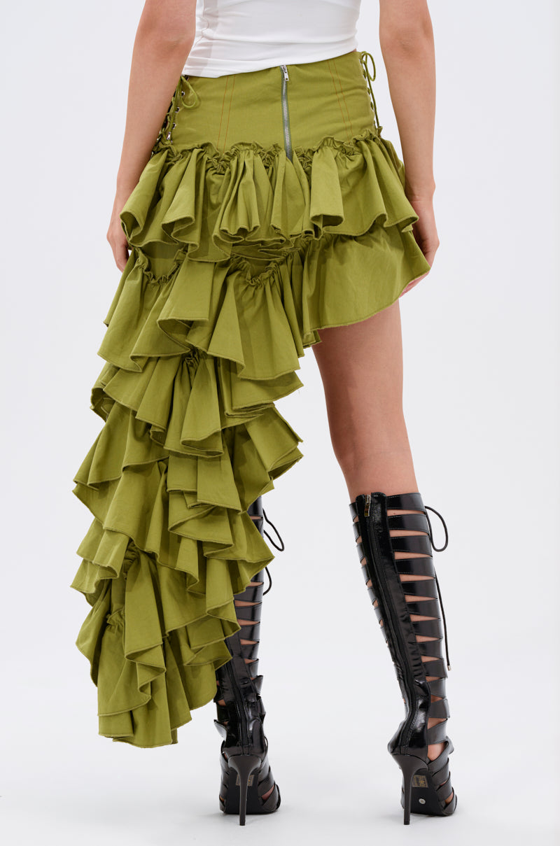 A person wears a white top, knee-high black lace-up boots, and the CLAIRE HIGH LOW RUFFLE SKIRT—an asymmetrical olive green skirt with dramatic layered ruffles on one side and a back zipper.