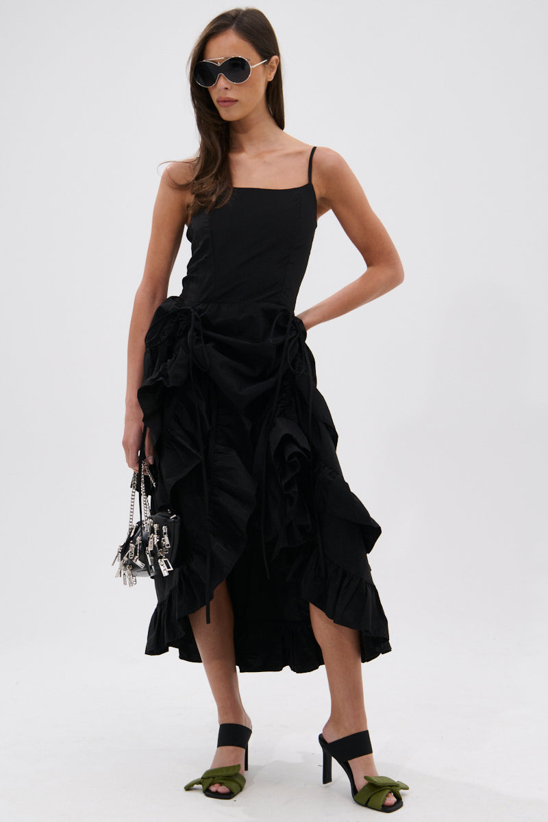 A woman stands confidently against a white background, wearing the ADDED DRAMA RUFFLED MIDI DRESS IN BLACK with adjustable spaghetti straps, sunglasses, green open-toe heels, and holding a small handbag.