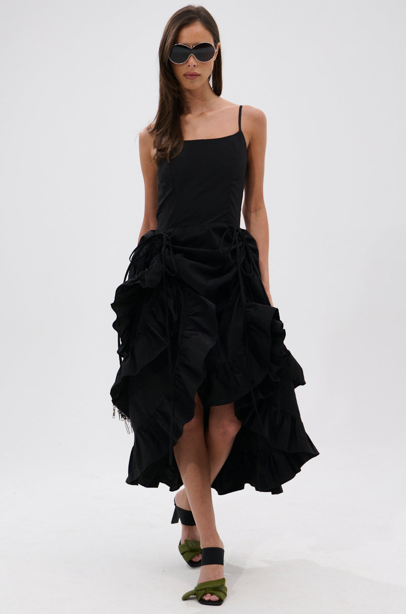A woman walks confidently against a white background wearing the ADDED DRAMA RUFFLED MIDI DRESS IN BLACK, featuring adjustable spaghetti straps and a ruffled high-low hem, paired with green open-toe heels and sunglasses.