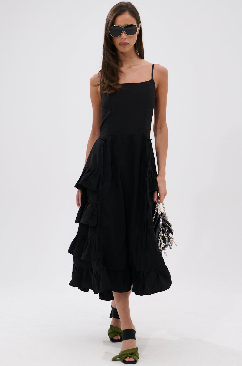 A woman in the ADDED DRAMA RUFFLED MIDI DRESS IN BLACK with adjustable straps, dark sunglasses, green open-toe sandals, and a black-and-white patterned handbag stands against a white background.