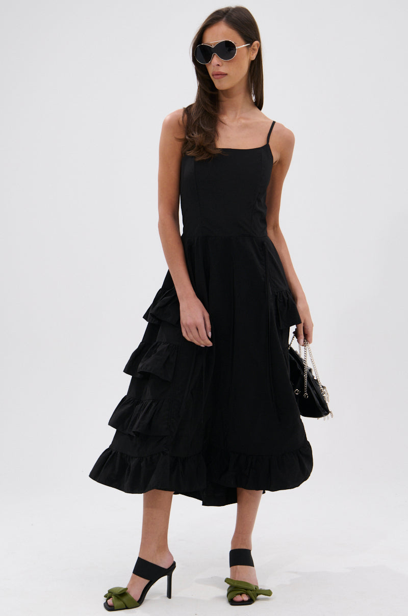 A woman stands against a plain background wearing the ADDED DRAMA RUFFLED MIDI DRESS IN BLACK with adjustable spaghetti straps and ruffles, paired with sunglasses, a black chain strap bag, and green heeled sandals.