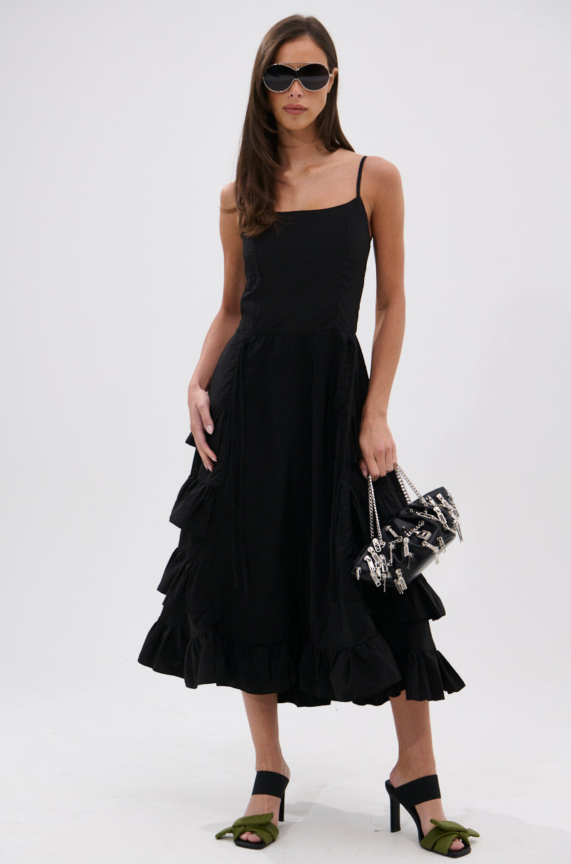 A woman wears the ADDED DRAMA RUFFLED MIDI DRESS IN BLACK, black heels with green bows, dark sunglasses, and carries a black handbag with silver accents while standing against a plain white background.