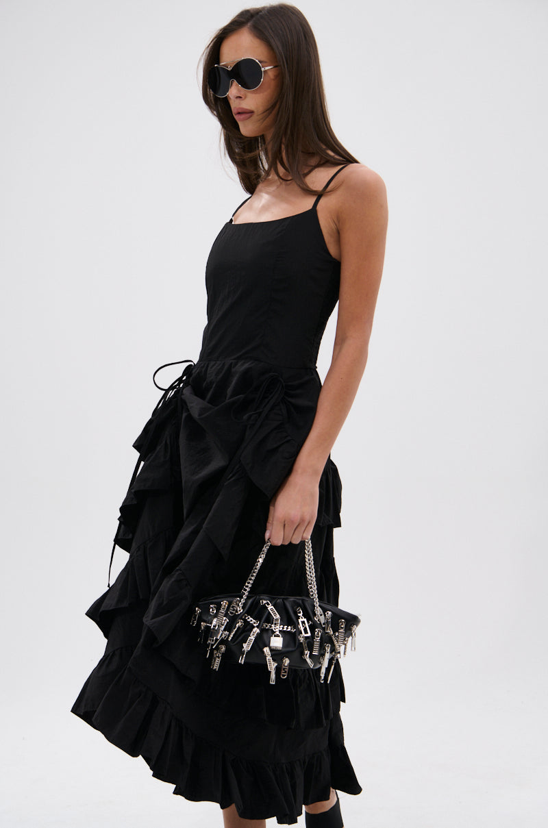 A woman with long brown hair wears sunglasses and the ADDED DRAMA RUFFLED MIDI DRESS IN BLACK, which features an asymmetrical high-low skirt. She holds a black bag with silver key charms, standing against a plain white background.