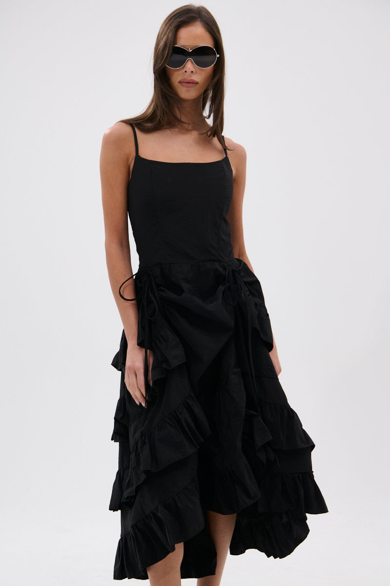 A woman in dark sunglasses wears the ADDED DRAMA RUFFLED MIDI DRESS IN BLACK, featuring adjustable spaghetti straps and a sleeveless ruffled design, standing against a plain light background.